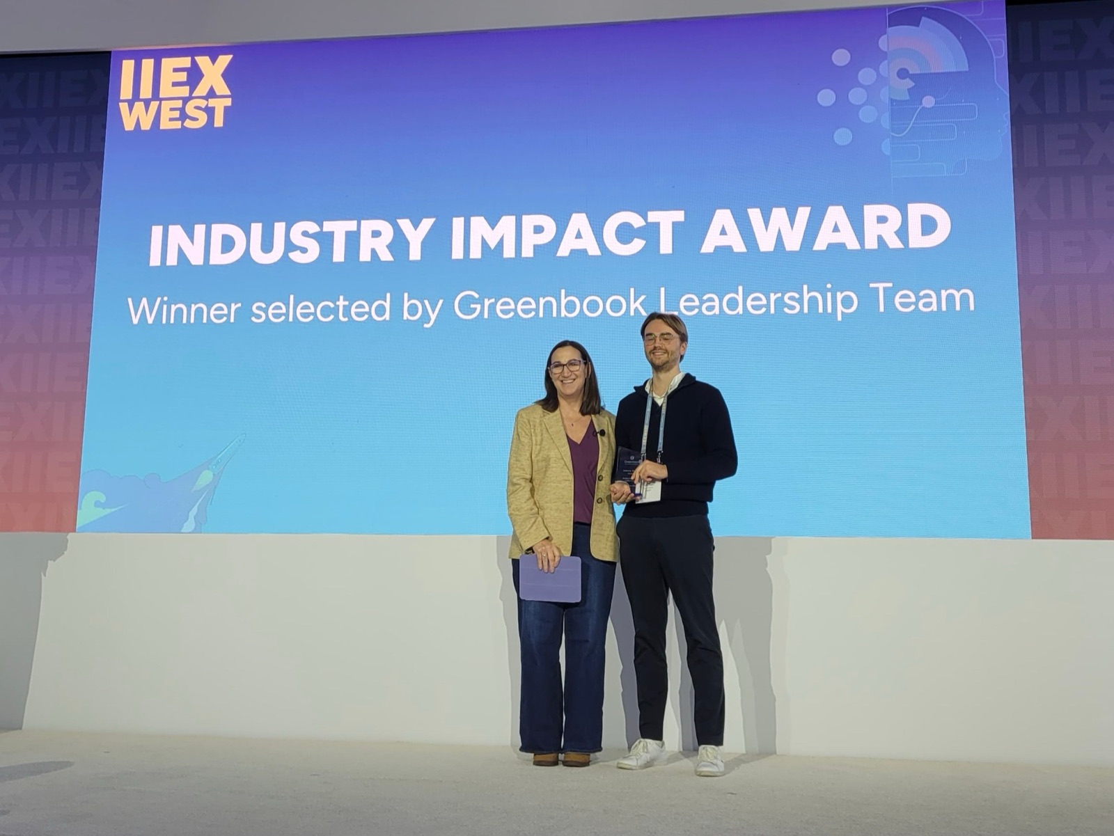Adrien Vermeirsch holding the Industry Impact Award on stage at IIEX West, next to the presenter, under a screen reading: Industry Impact Award, Winner selected by Greenbook Leadership Team